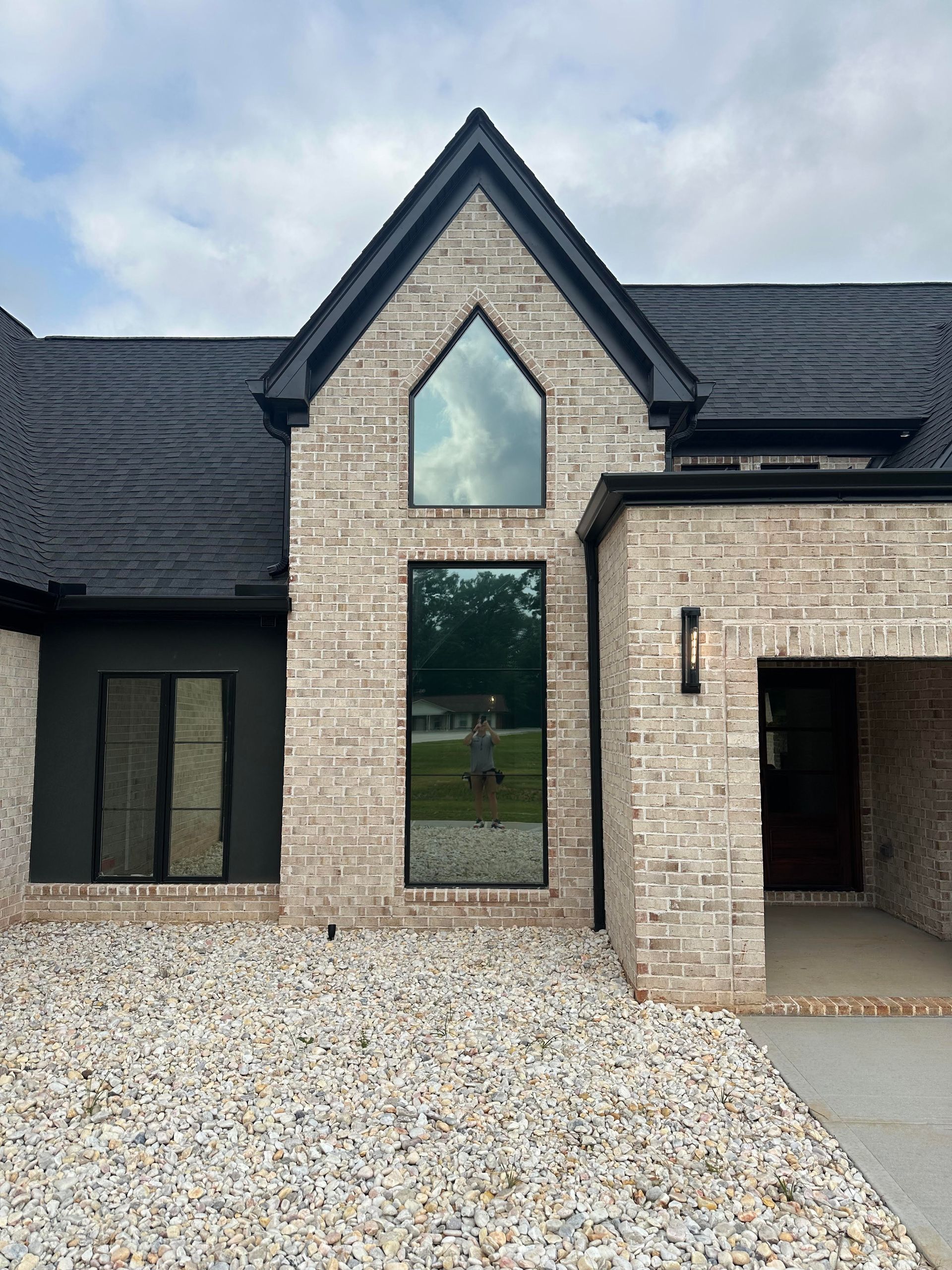 A modern house exterior with beige brick, a steep peaked roof, dark trim, and a central floor-to-ceiling window feature.