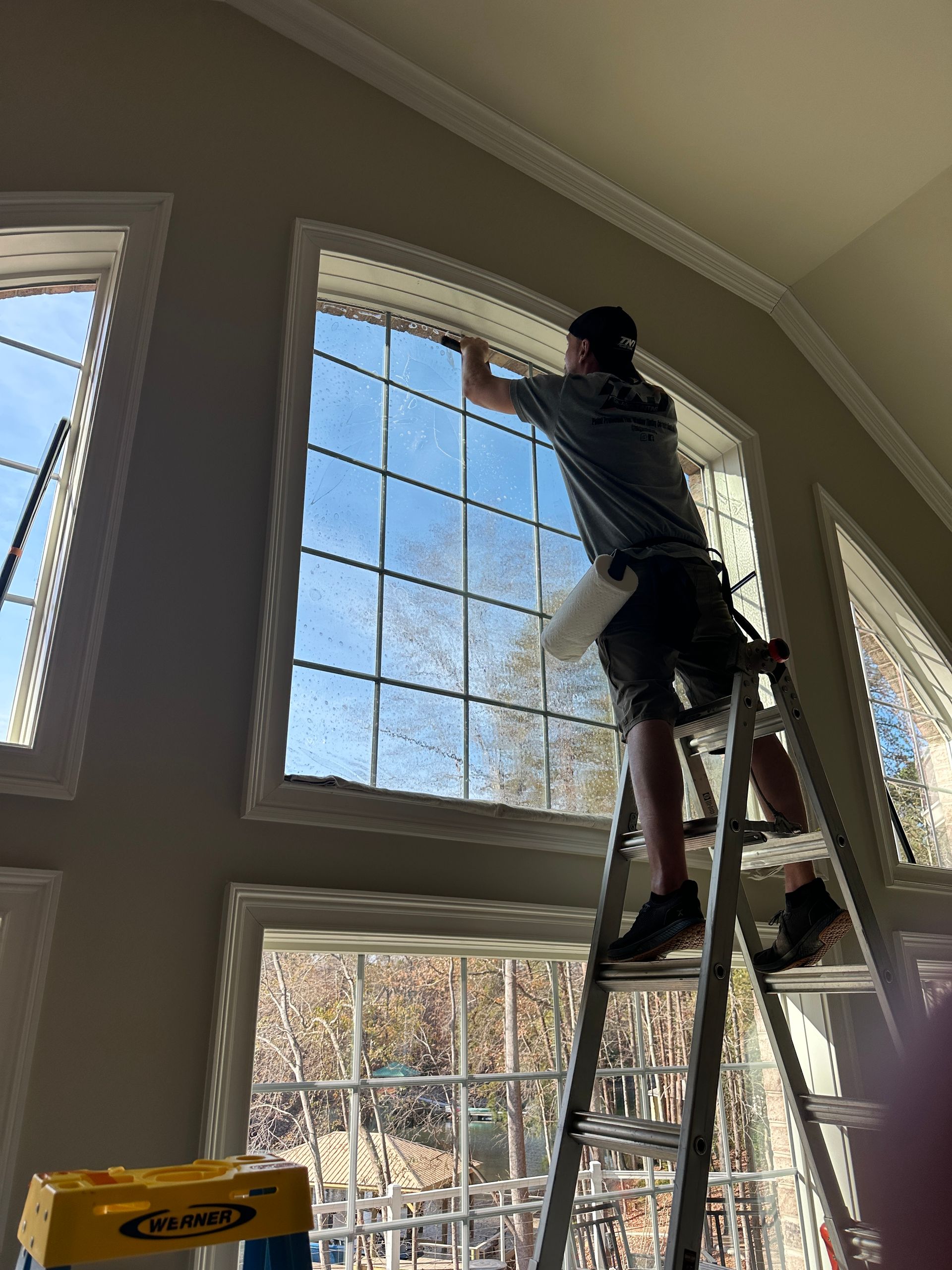 A person stands on a ladder cleaning the upper portion of a large, multi-pane window inside a home.