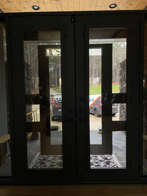 Black double entry doors with rectangular glass panels, seen from inside, showing a driveway and parked van outside.