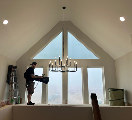 A worker installs large glass window panels in a room overlooking a lake, with two ladders present.