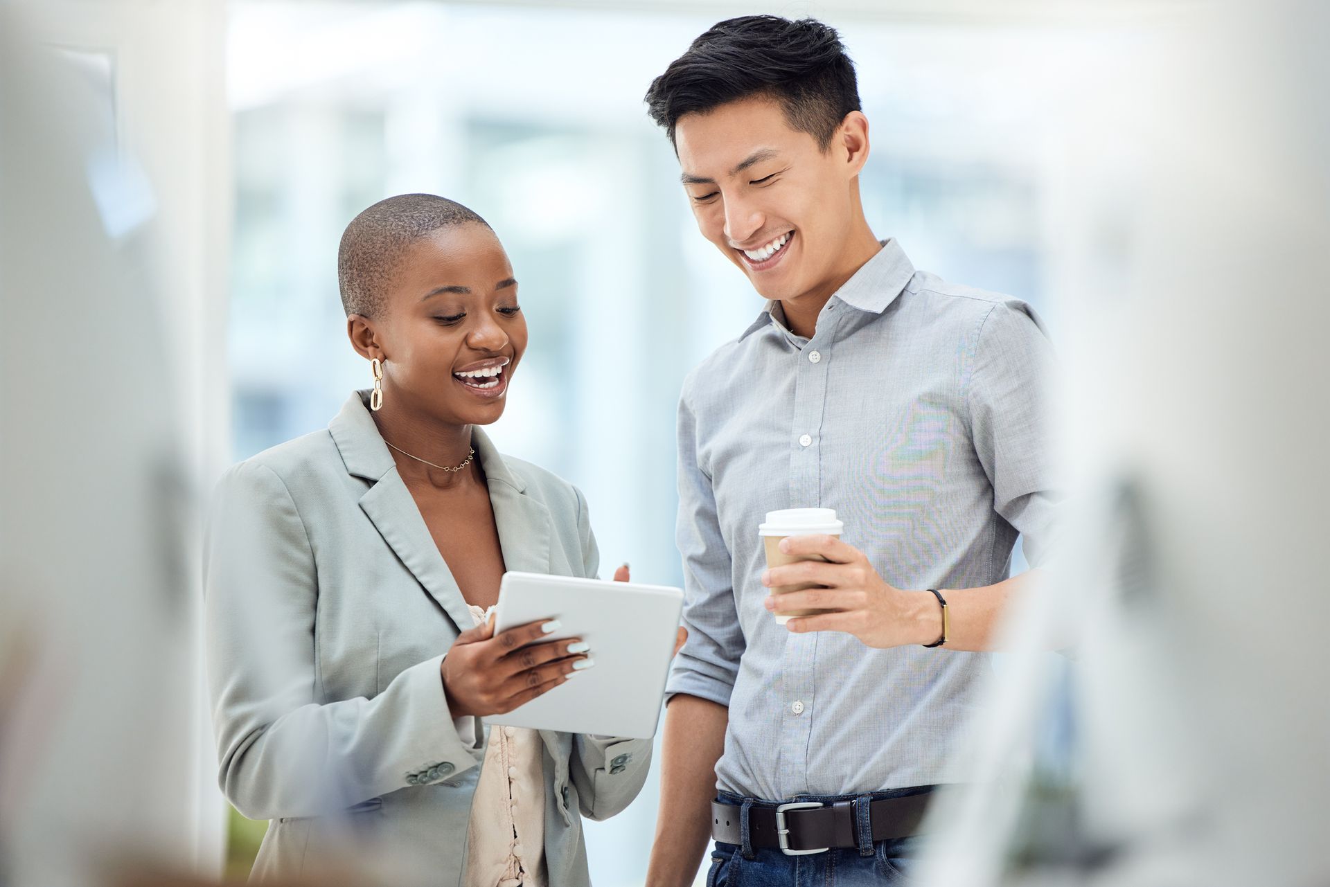 Woman showing tablet to man, both smiling, holding coffee. Indoors, bright light.