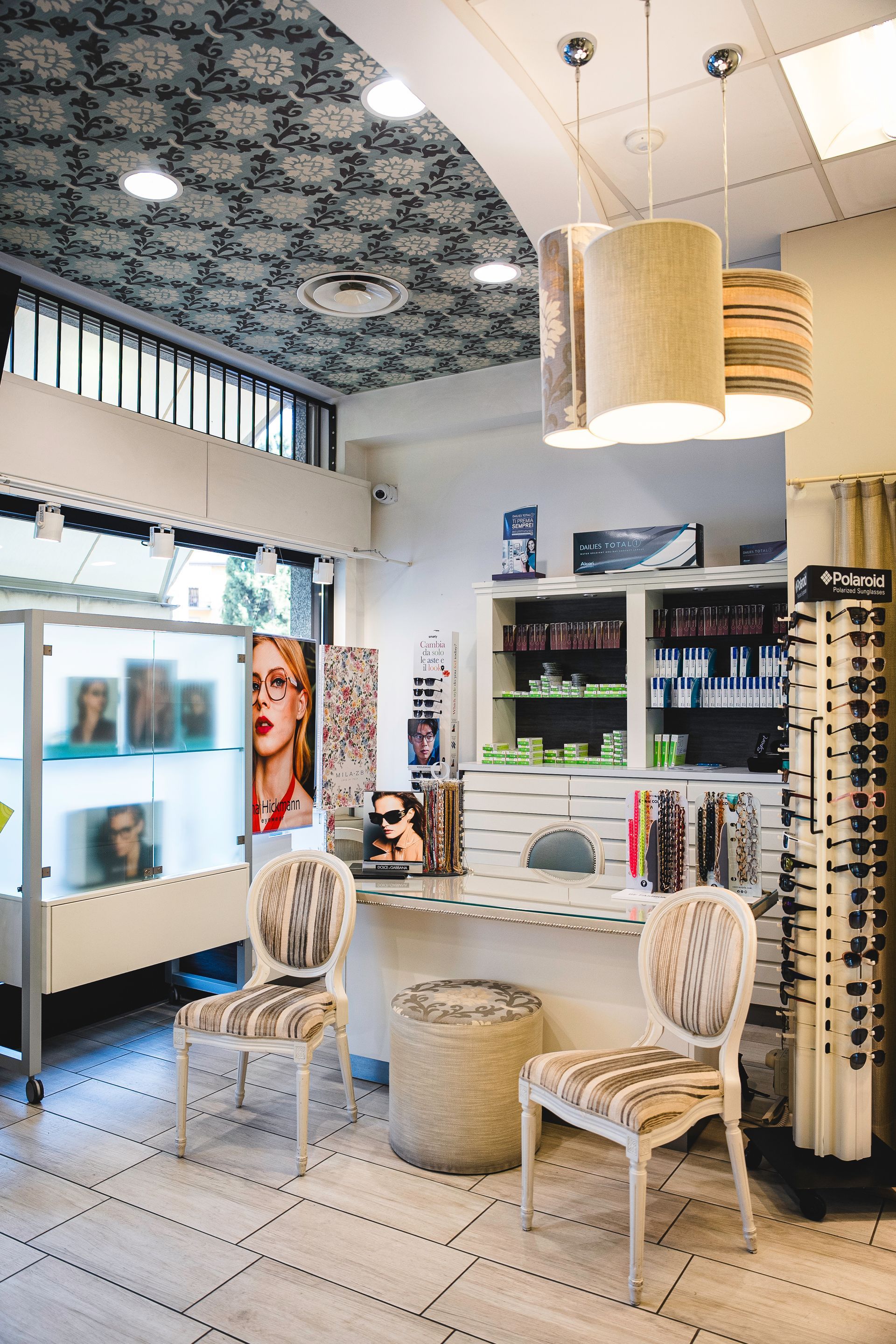 Interno dello studio di un ottico con sedie, bancone, espositori per occhiali e lampade a sospensione.