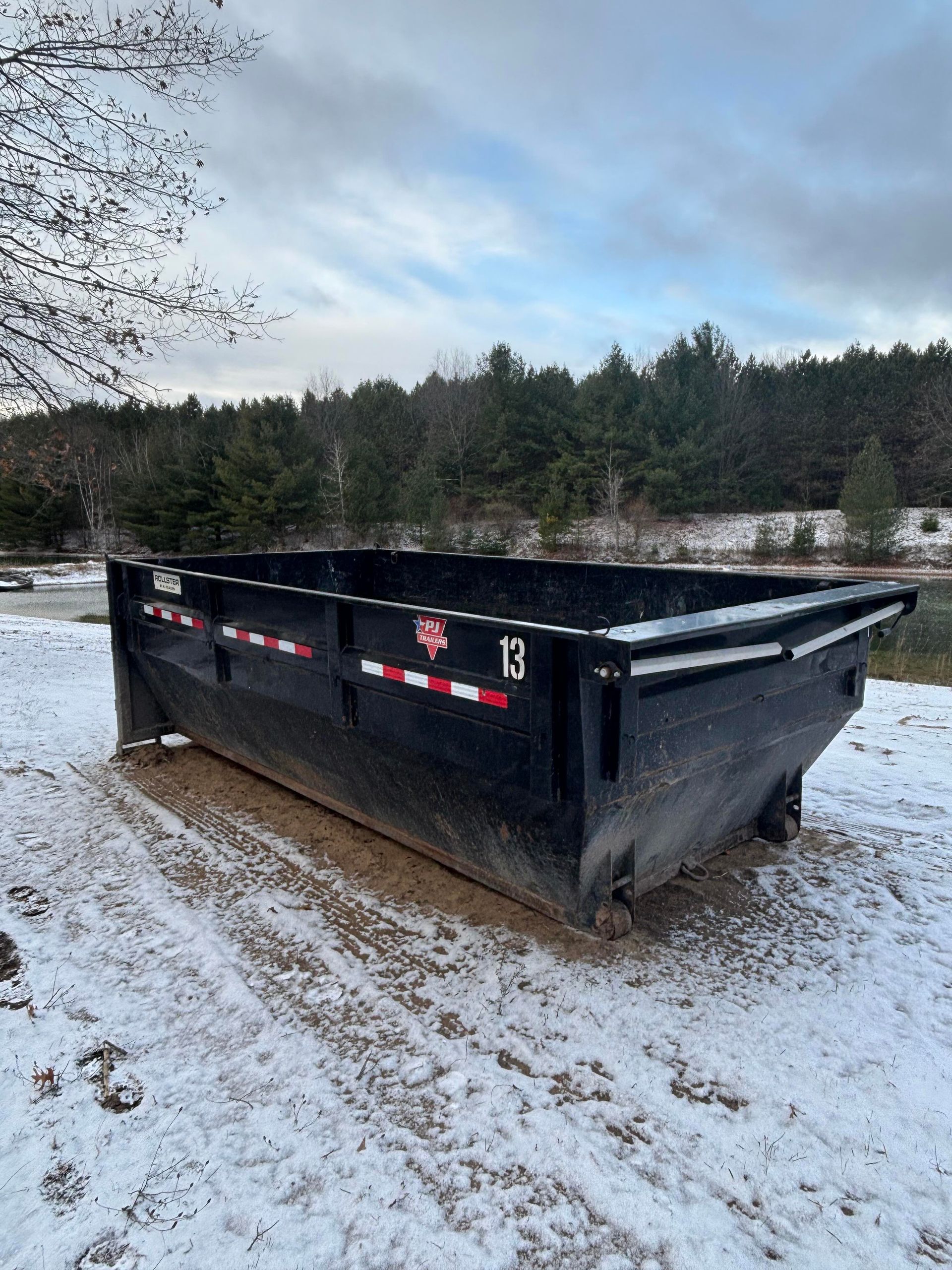 Black dumpster in a snowy outdoor setting with trees in the background.