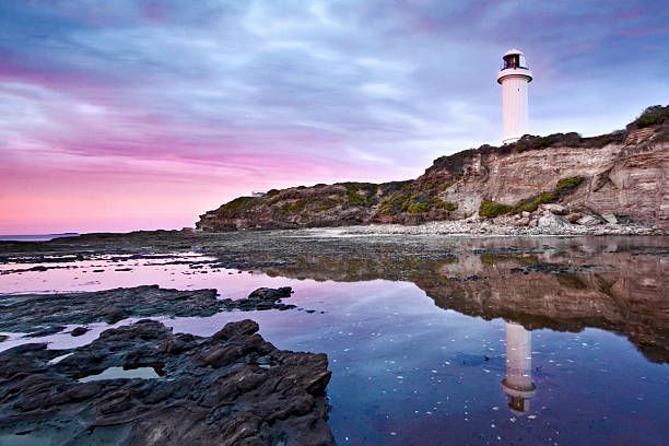A Lighthouse Is Reflected In The Water Near A Rocky Shoreline — Bastion Electrical In Wollongong, NSW
