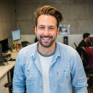 Smiling man in light blue shirt, white tee, indoors, office setting with computers, other people in background.