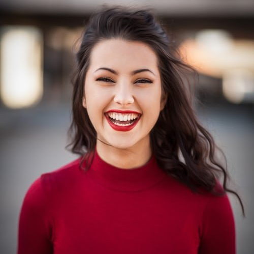 Woman with dark hair, wearing a red turtleneck, smiling broadly, outdoors.