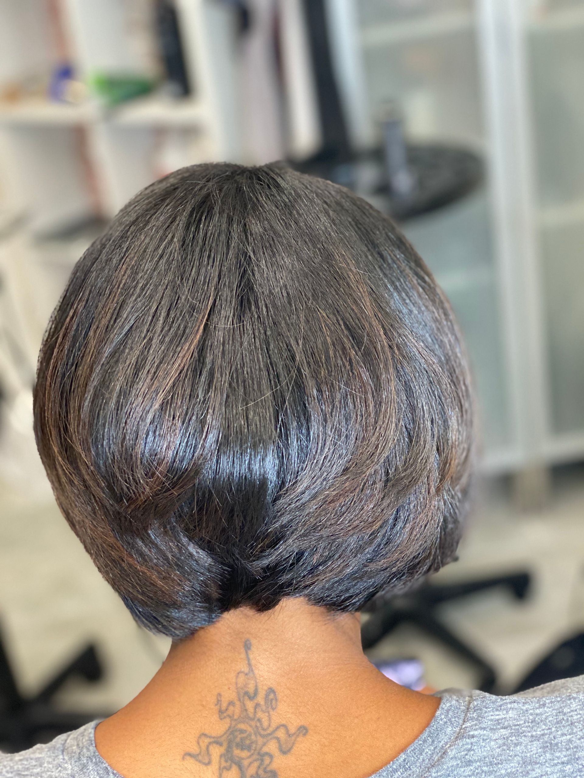 Woman with a short, layered, dark bob haircut, showing a tattoo on her neck, in a salon setting.
