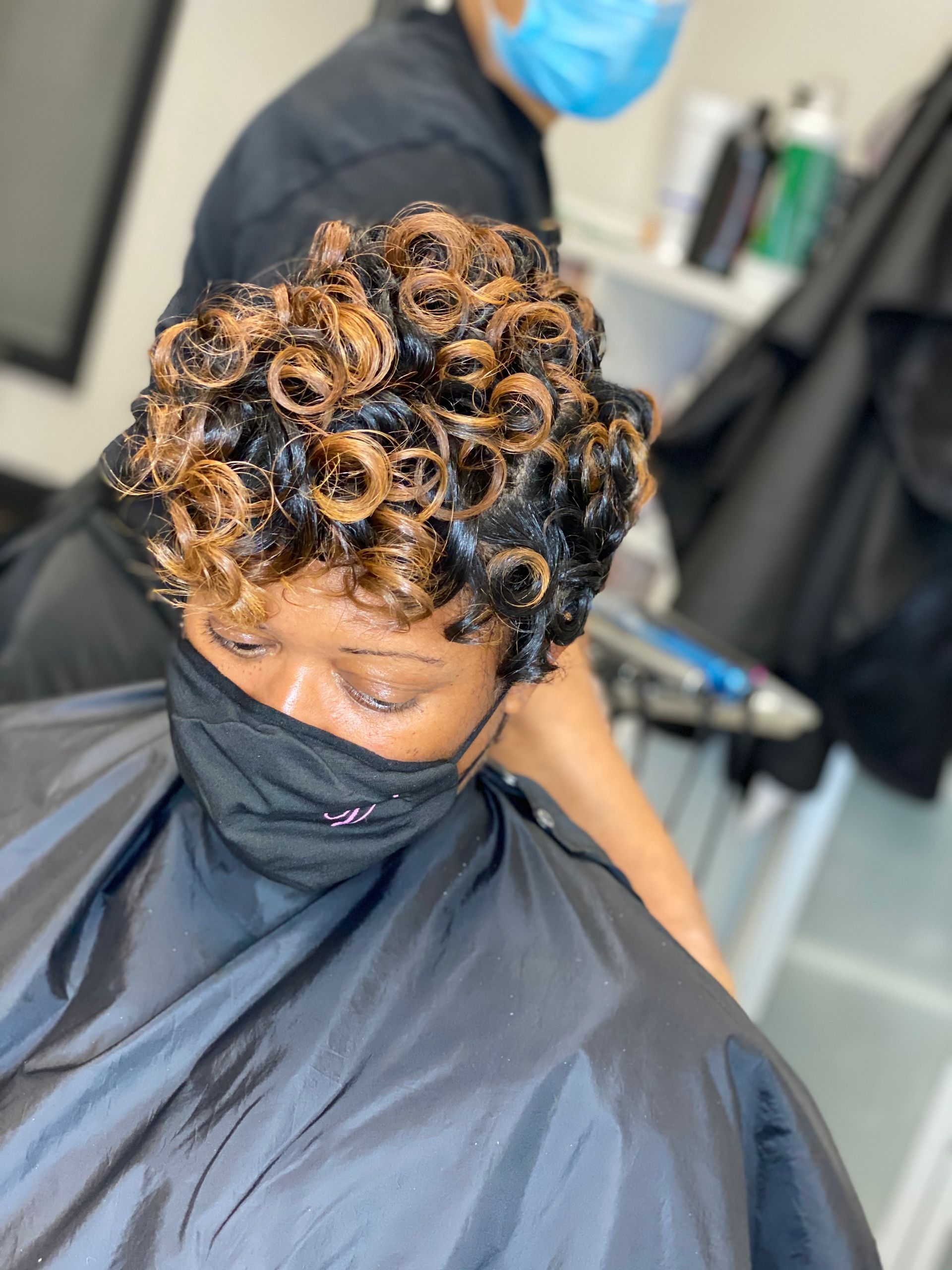 Woman with short, curled hair getting styled by a person in a salon, both wearing face masks.