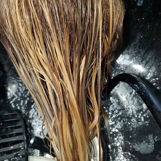Woman with long dark hair touching her head, back to the viewer. Beige top, plain background.