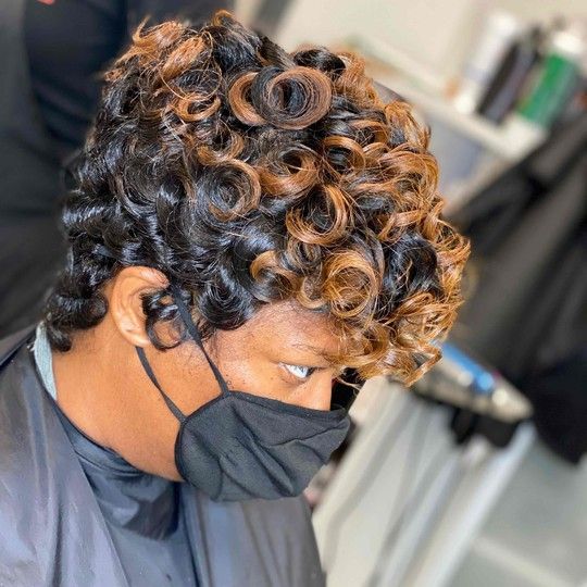 Woman with black hair styled with curled honey-blonde highlights, wearing a black mask and cape.