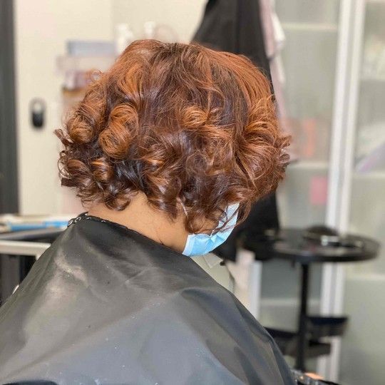 Woman with curly, auburn hair in a salon, wearing a mask and a black cape.