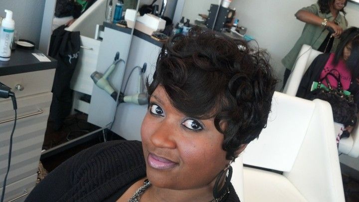 Woman with dark hair, smiling at a salon. Styling products and hair dryers in background.