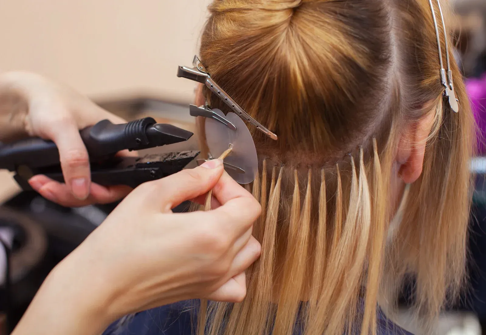 Hands attaching hair extensions to blonde hair with a heated tool in a salon.