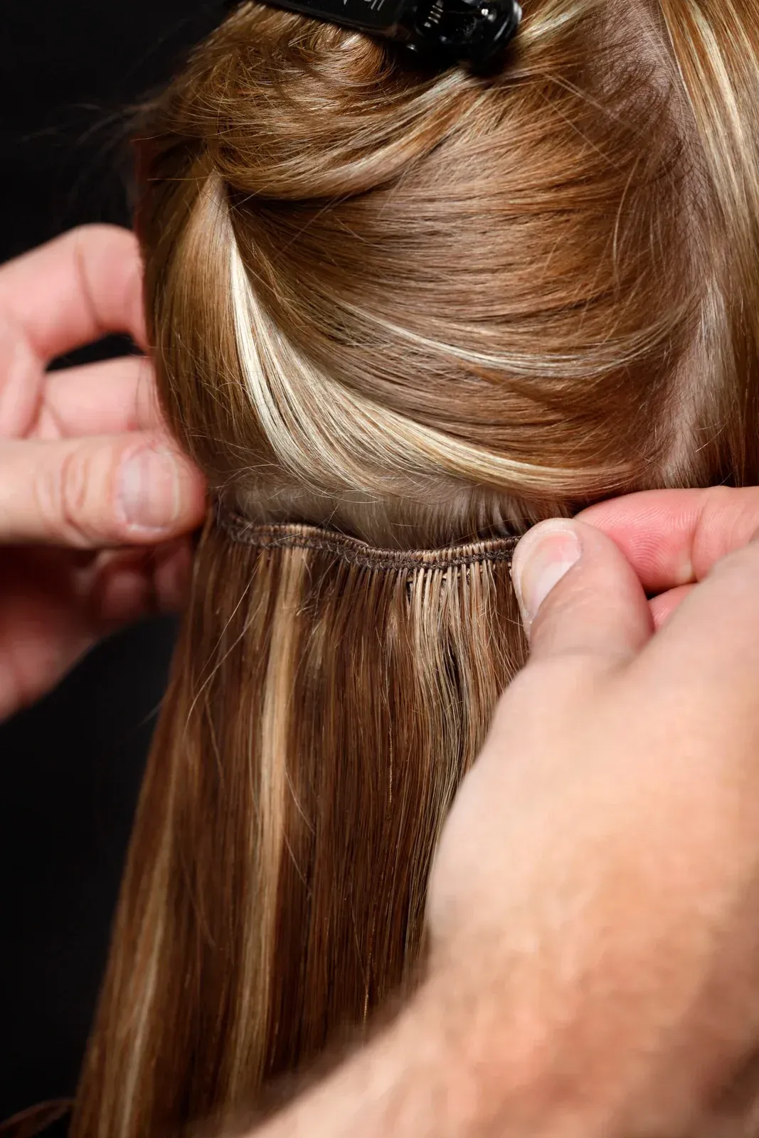 Hands attaching hair extensions to a person's natural hair; blonde and brown strands.