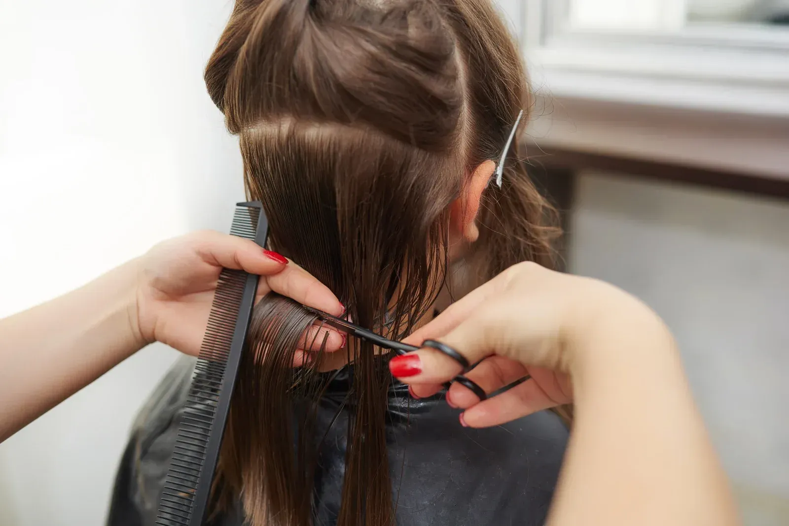 Hands cutting a person's long brown hair with scissors and a comb in a salon setting.