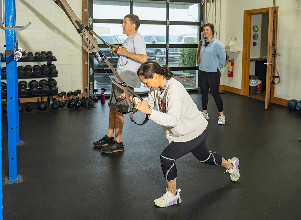 A woman in a red shirt is squatting in a gym while a man stands behind her.
