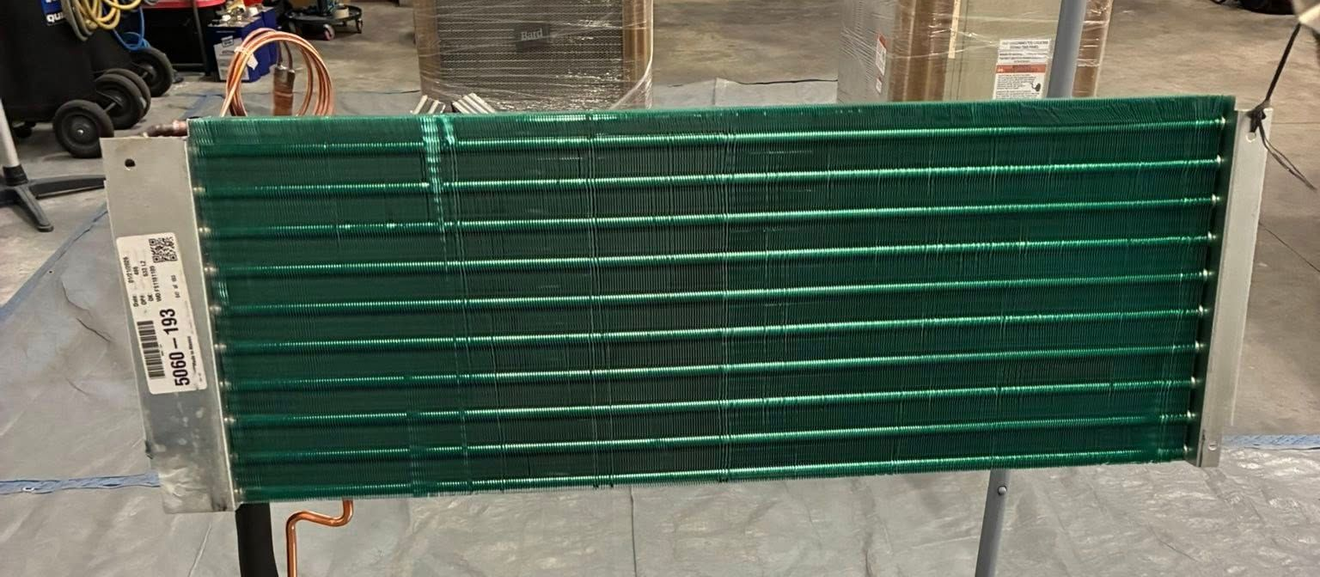 An HVAC evaporator coil with a green protective coating, mounted on metal side brackets in a workshop setting.