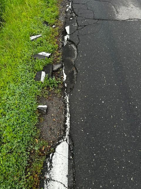 A road edge with crumbling asphalt and broken chunks of a white painted line bordering a grassy shoulder.