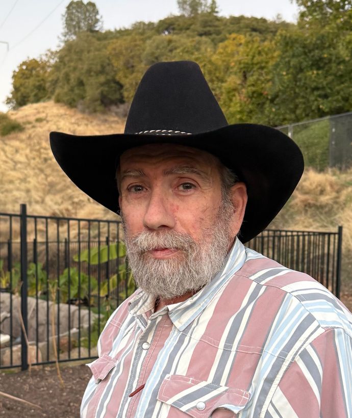A person wearing a black cowboy hat and a striped collared shirt outdoors with a fence and hillside in the background.