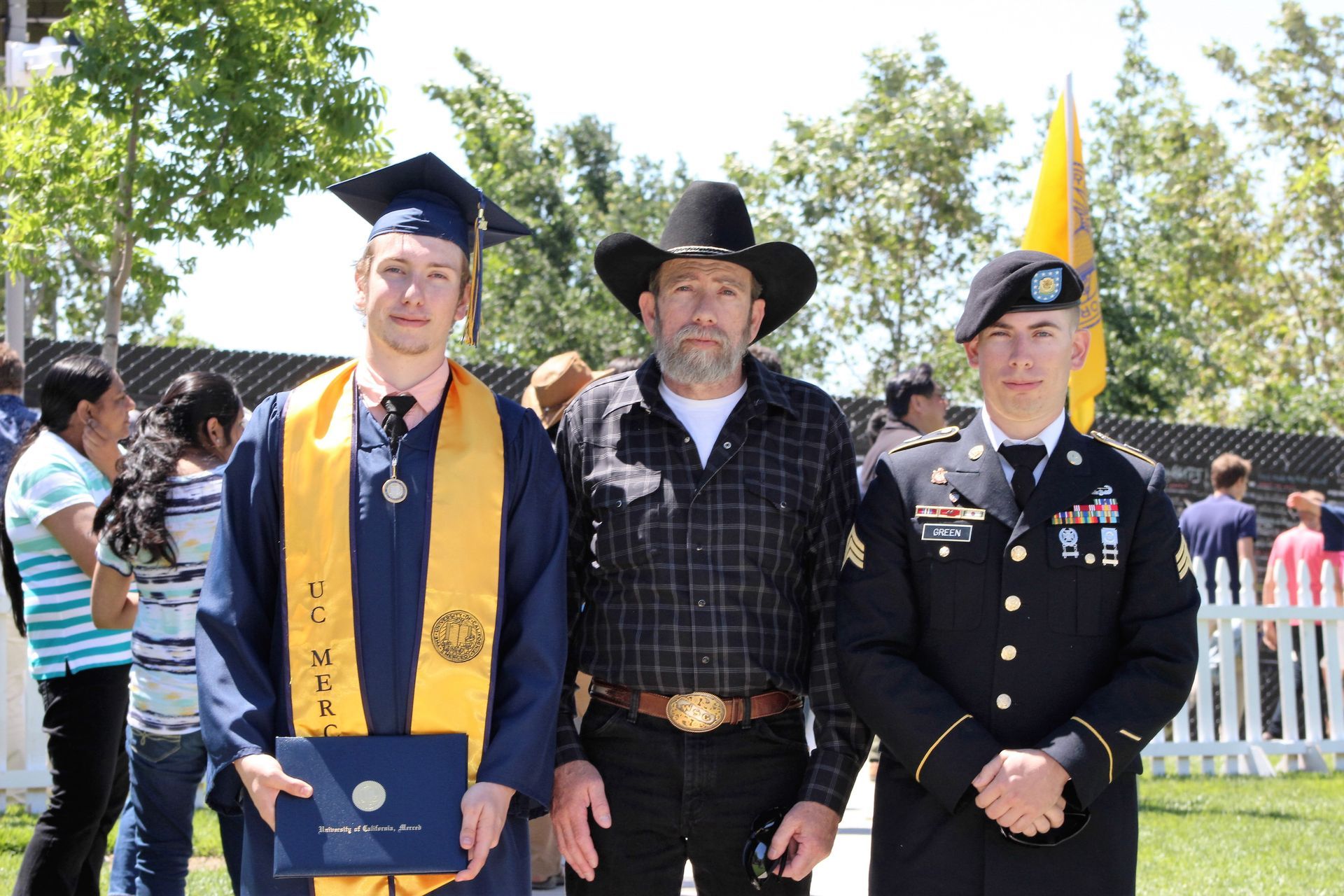 Three people pose outdoors: a student in graduation regalia, a person in a cowboy hat, and a soldier in uniform.