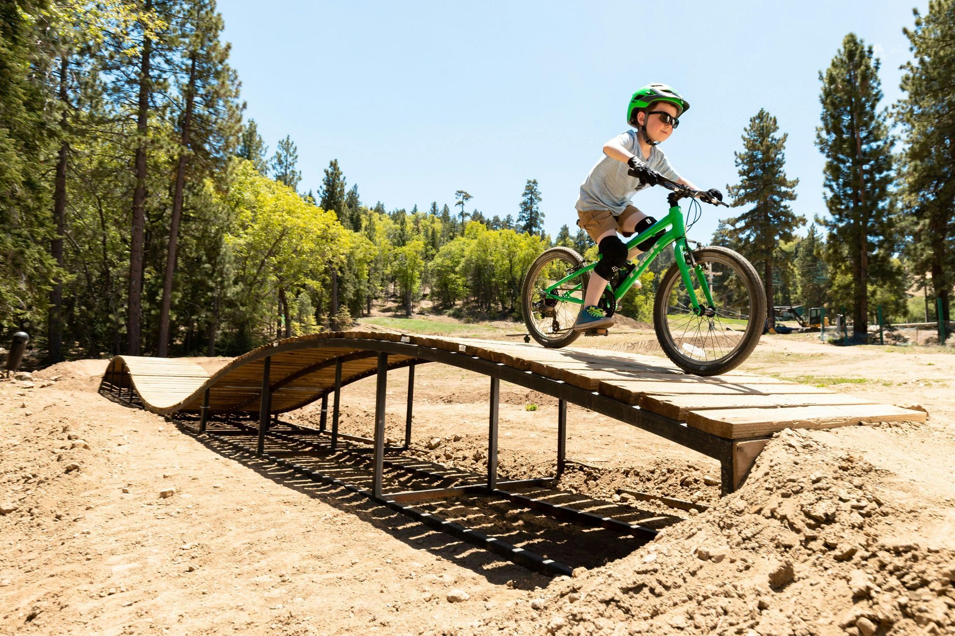 A child wearing a helmet rides a green mountain bike over a wooden roller bridge on an outdoor dirt track.