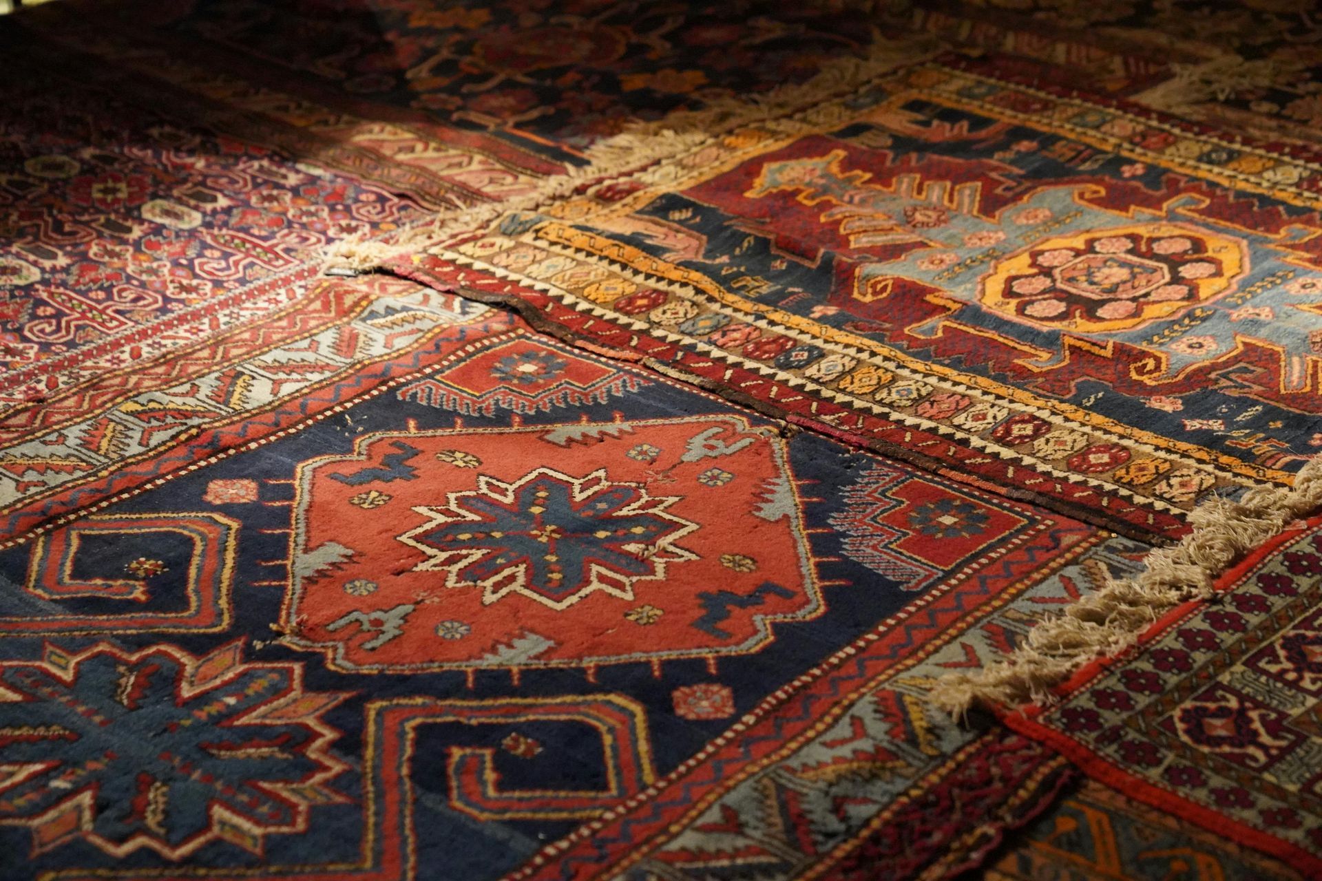 An assortment of overlapping traditional, ornate rugs in shades of red, blue, and gold with intricate geometric patterns.