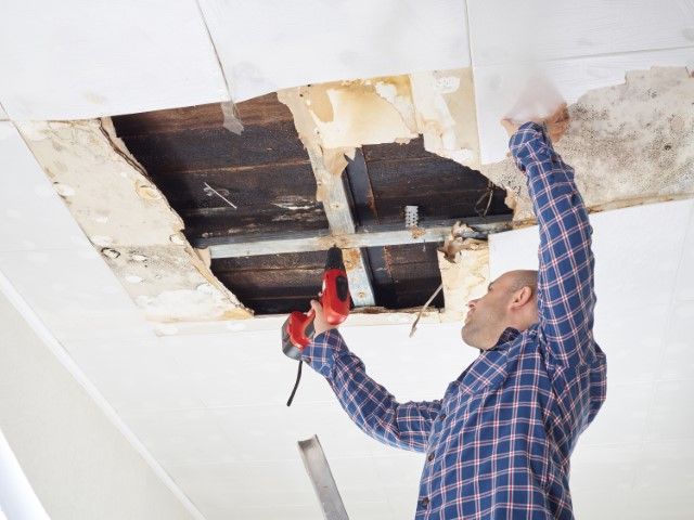 Worker repairing a damaged ceiling with exposed beams and drywall, using a red power drill.