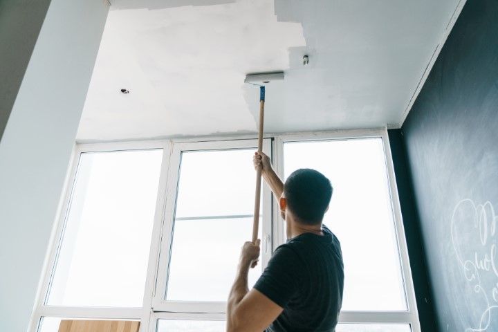 Person painting a white ceiling with a roller in a bright room