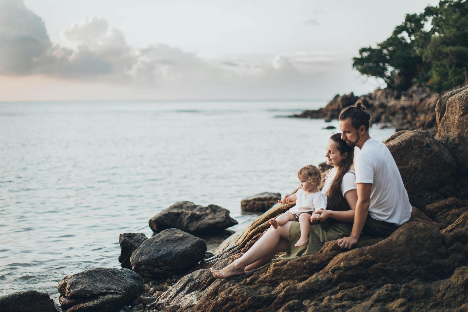 Family of three sits on rocks by the sea, looking out at the horizon.