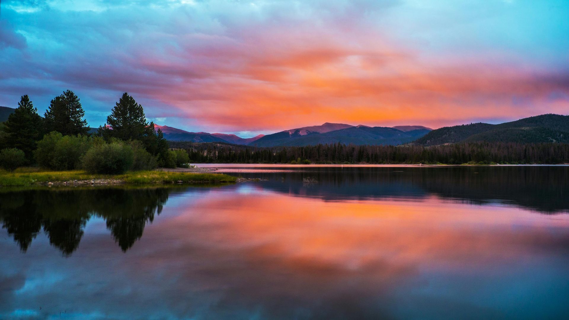 Sunset over a calm lake with reflections of orange and blue sky, surrounded by trees and mountains.