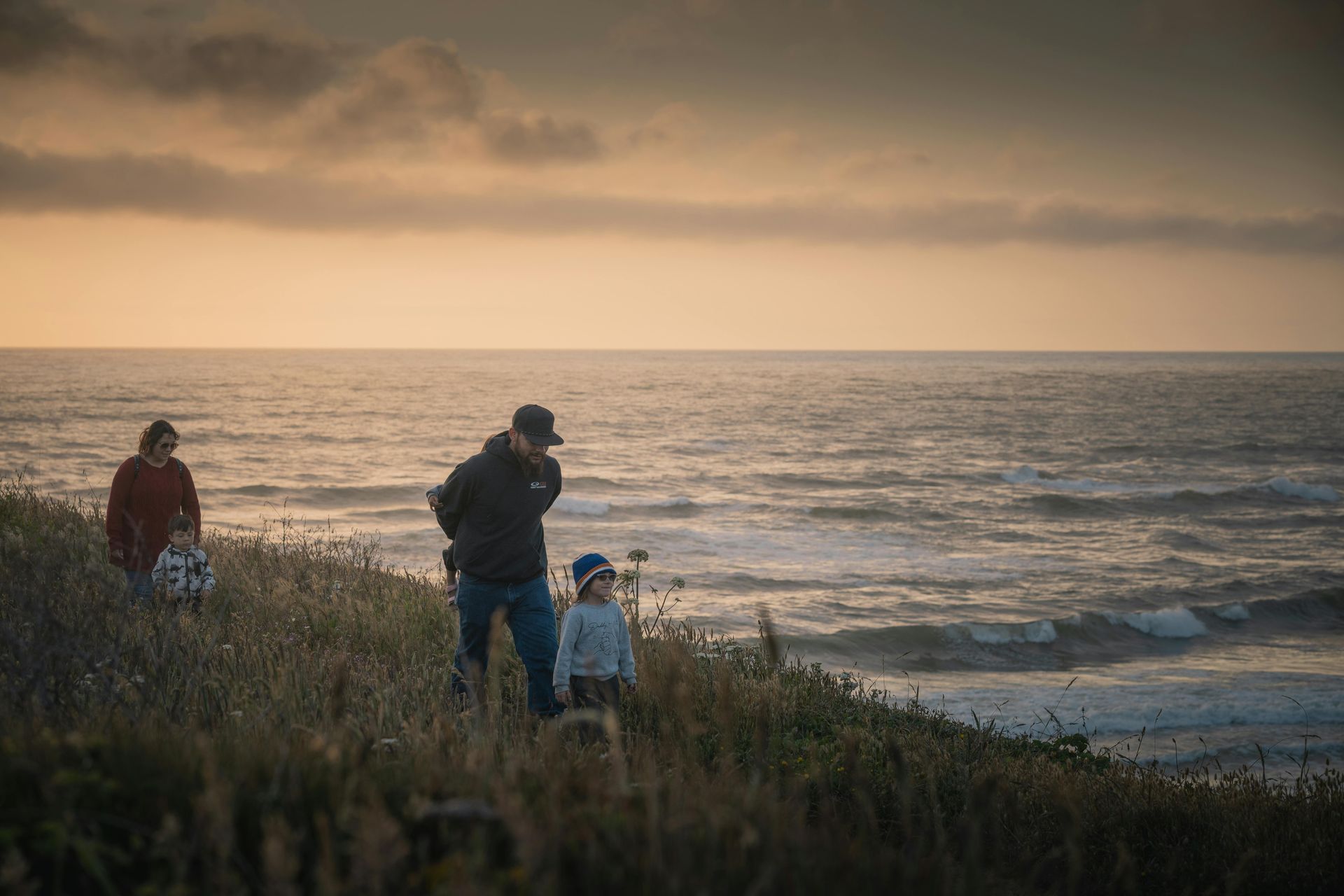 Family on a cliff overlooking the ocean at sunset; man, child, and woman, golden light.