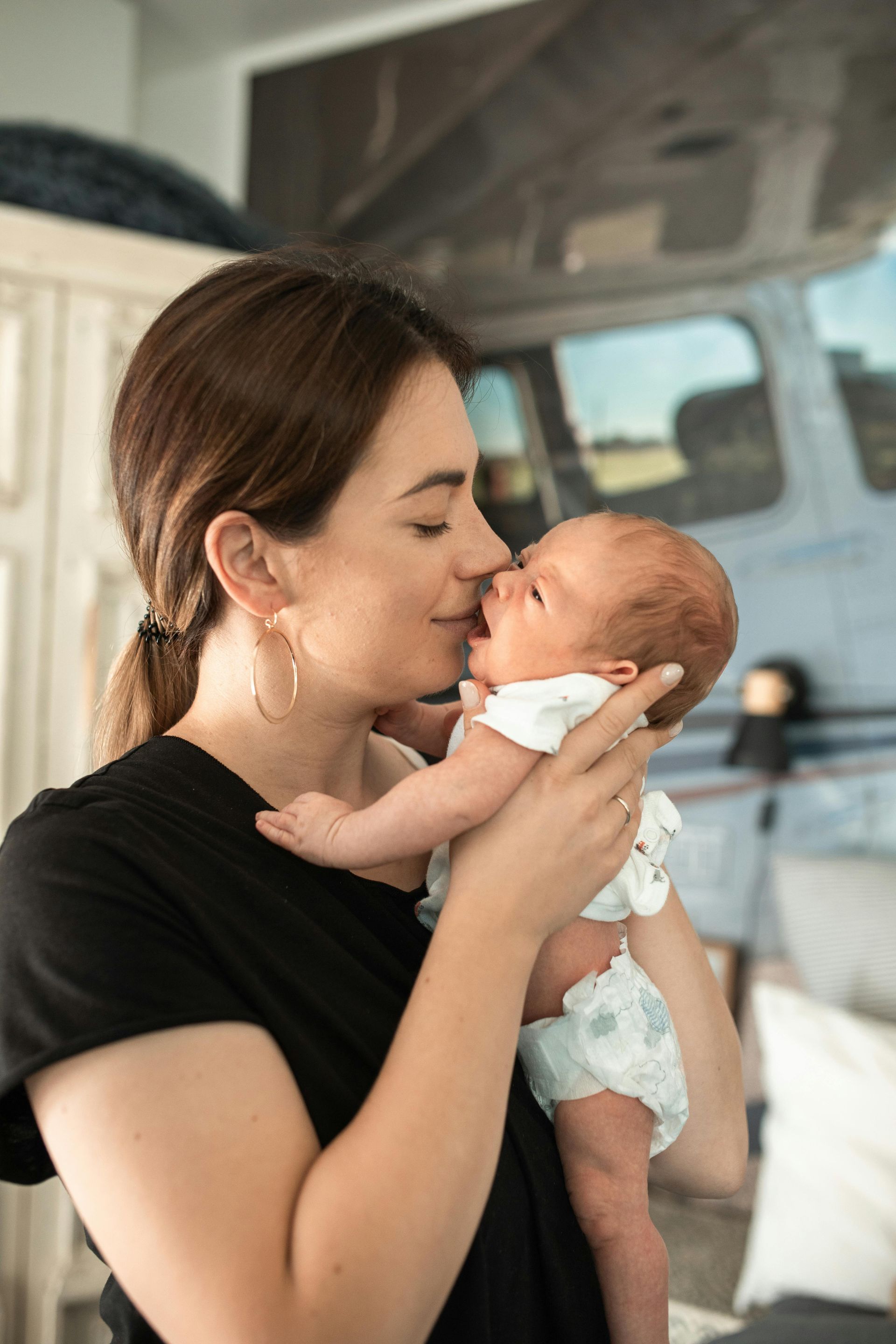 Woman in black shirt kisses a newborn baby's face. Inside, silver trailer visible in the background.