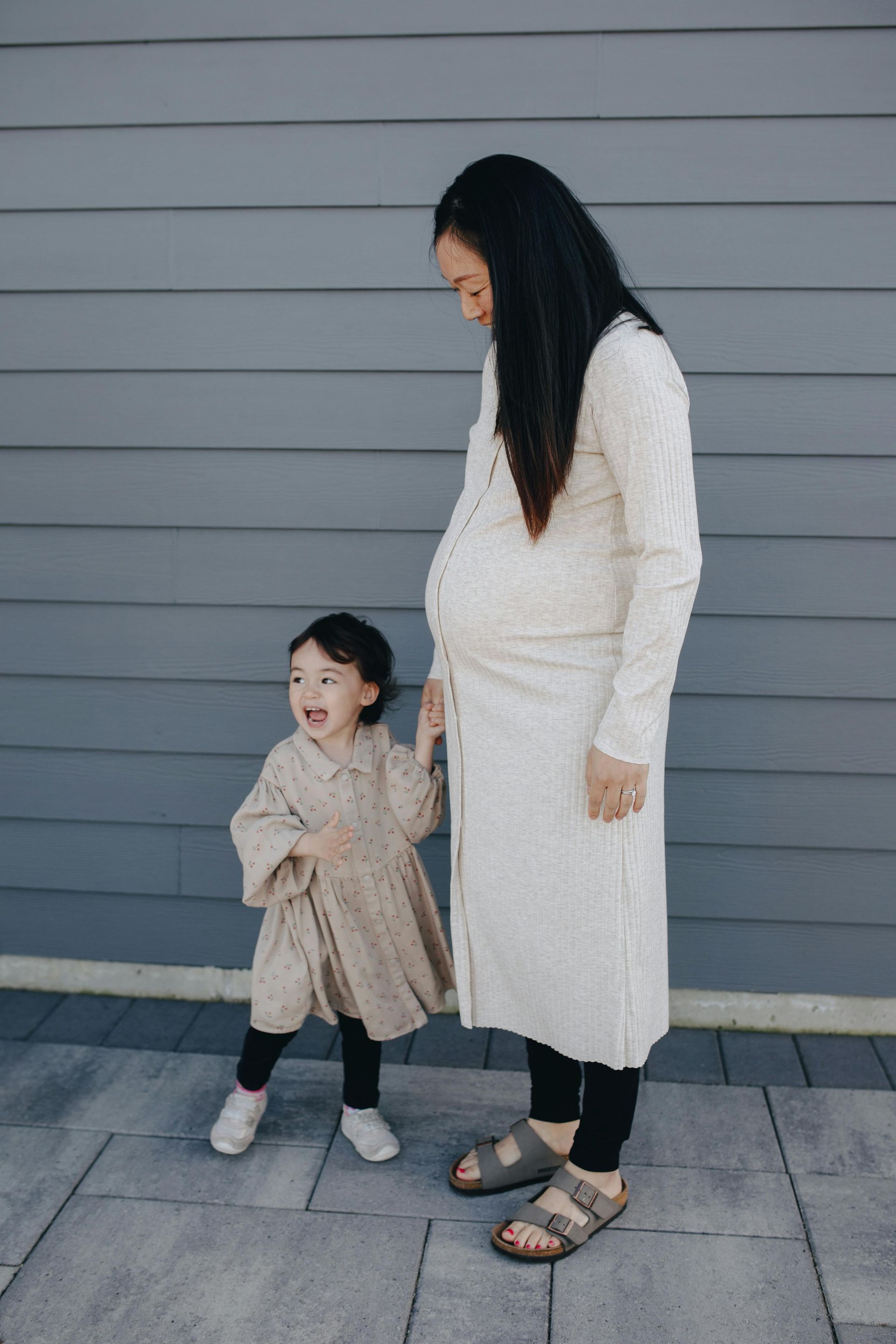 Pregnant woman and toddler stand in front of a grey wall. The toddler holds the woman's hand and looks up smiling.