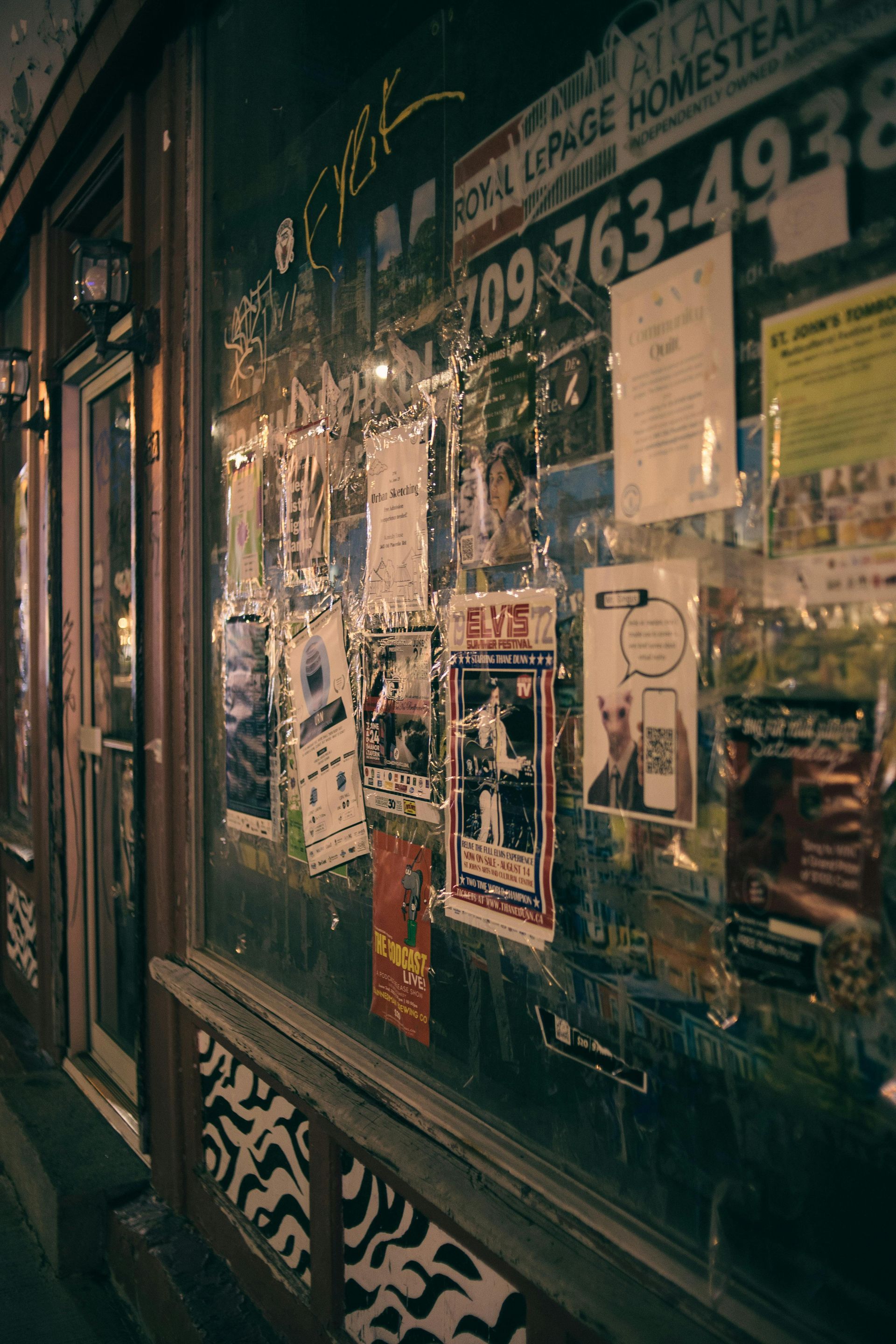 Exterior of a storefront window covered in posters, lit at night. Address visible: 709-763-4938.