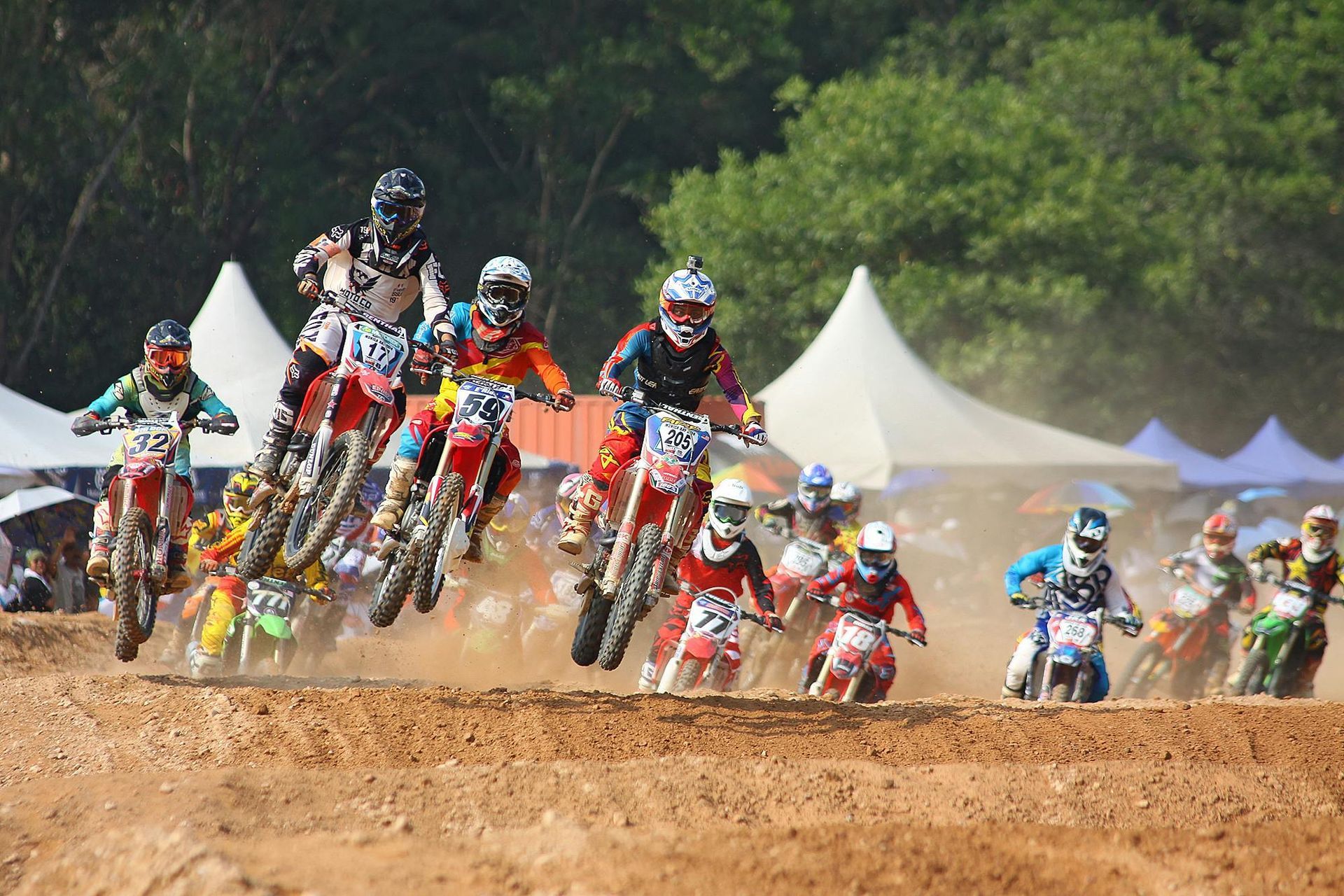 Motocross racers on dirt bikes in mid-air, racing on a dirt track; sunny day, spectators visible.