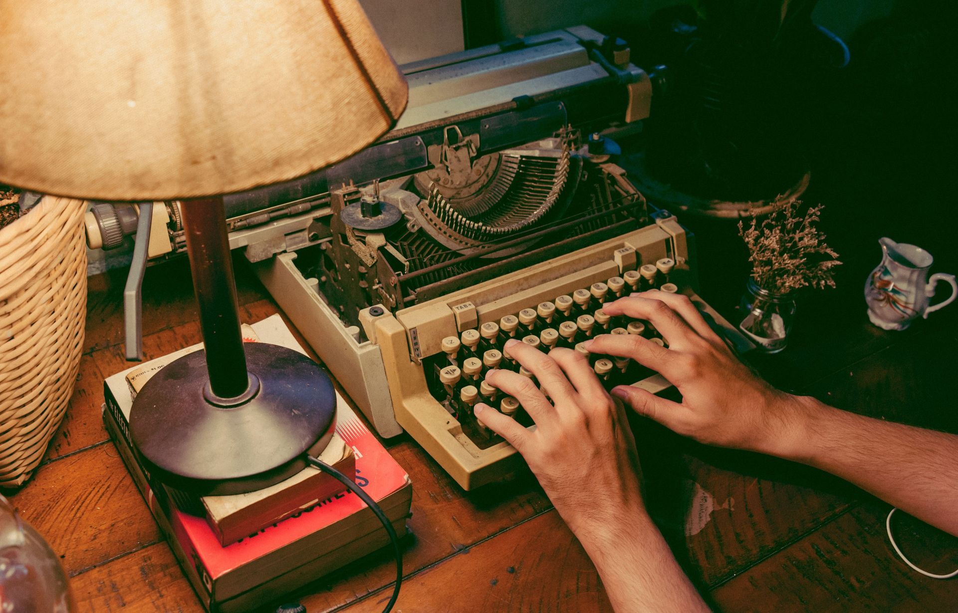 Hands typing on a vintage typewriter beside a lamp on a wooden desk.