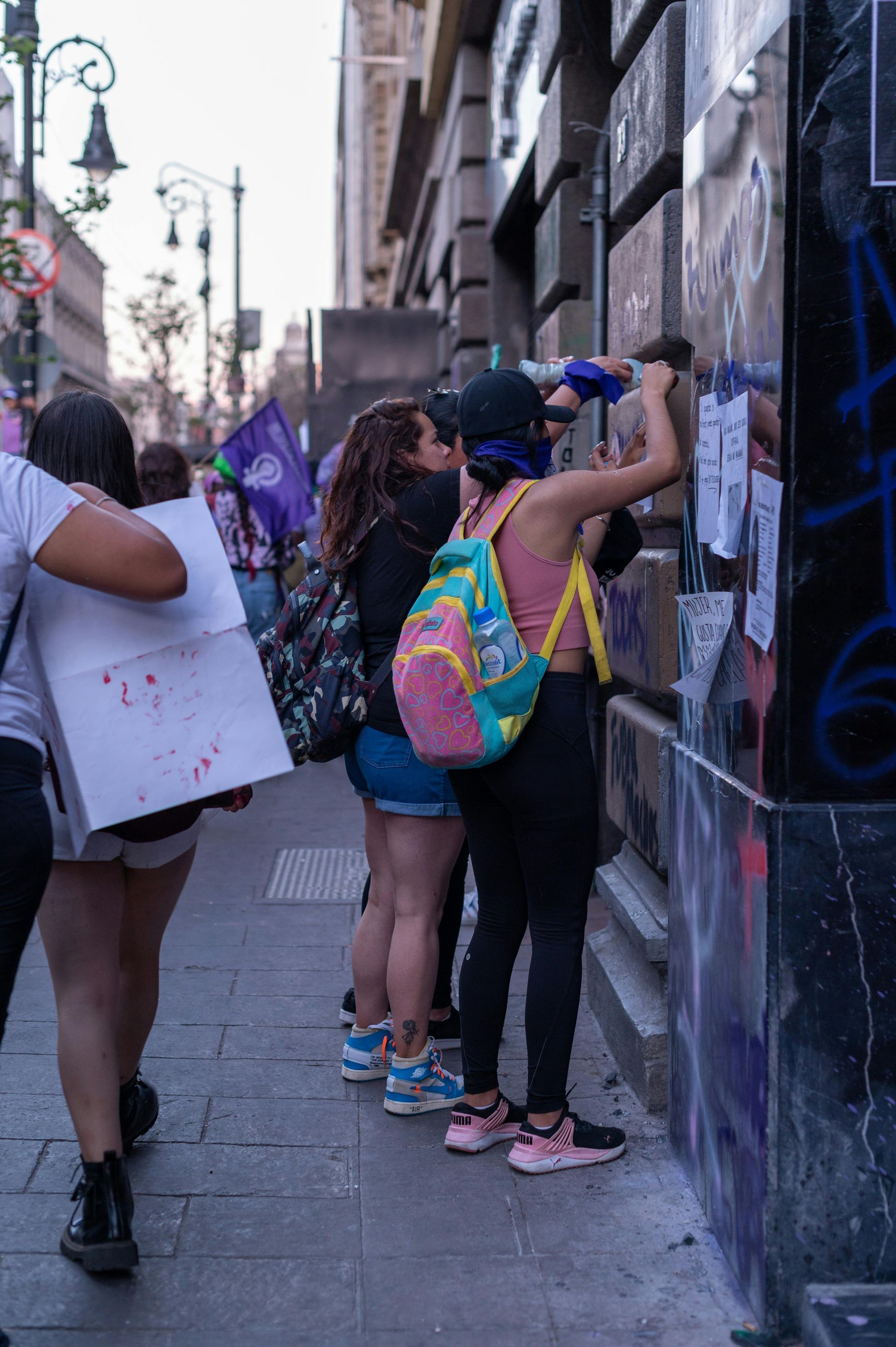 People spray-painting graffiti on a city wall, with backpacks and art supplies on a sidewalk.