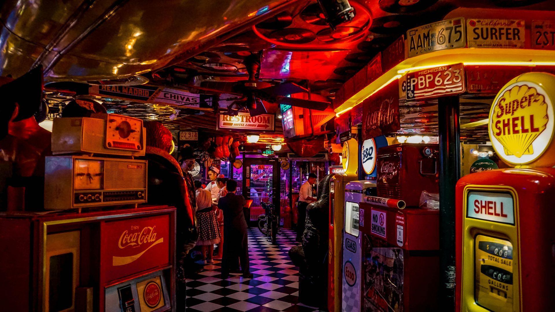 Interior of a diner with vintage signs, gas pumps, and a checkered floor. Dim lighting, red and gold accents.