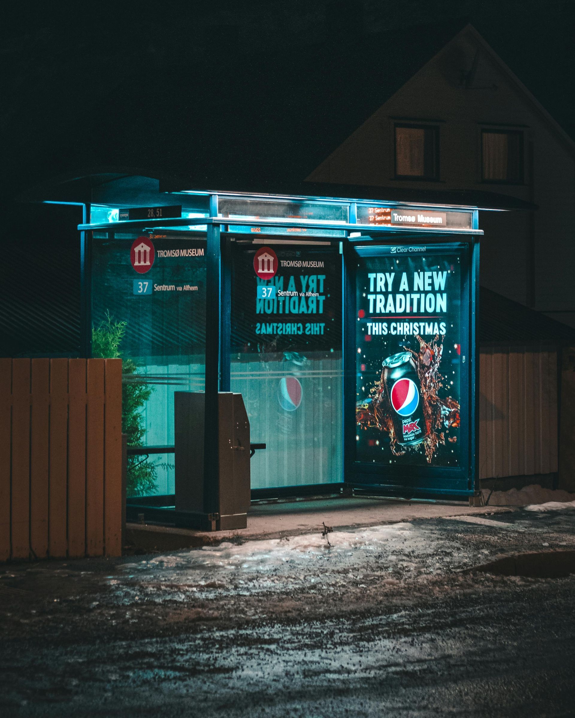 A brightly lit bus stop at night, advertising Pepsi. The shelter glows with blue lights, reflecting on the wet pavement.