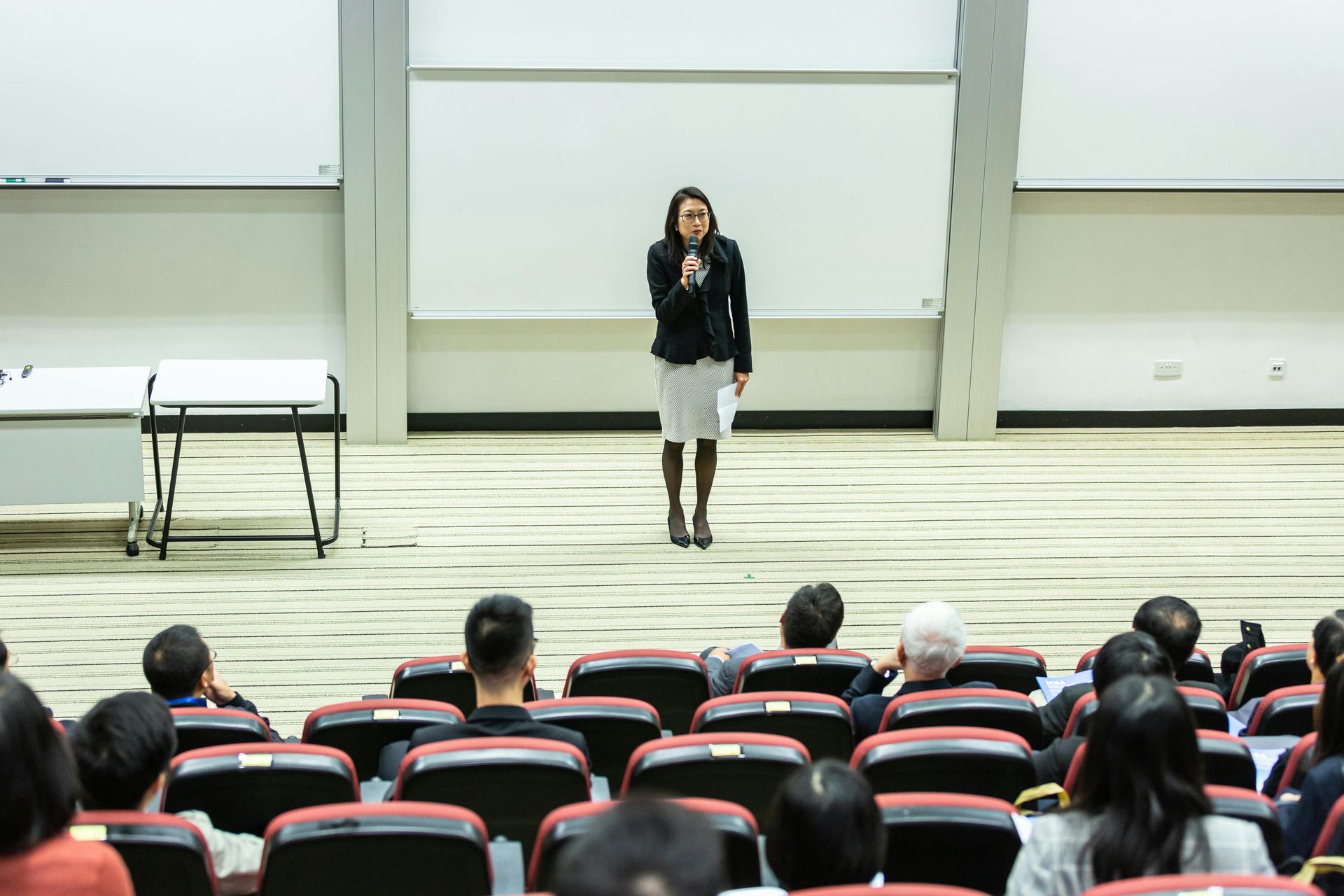 Woman speaking into a microphone in front of an audience, likely giving a presentation in a lecture hall.