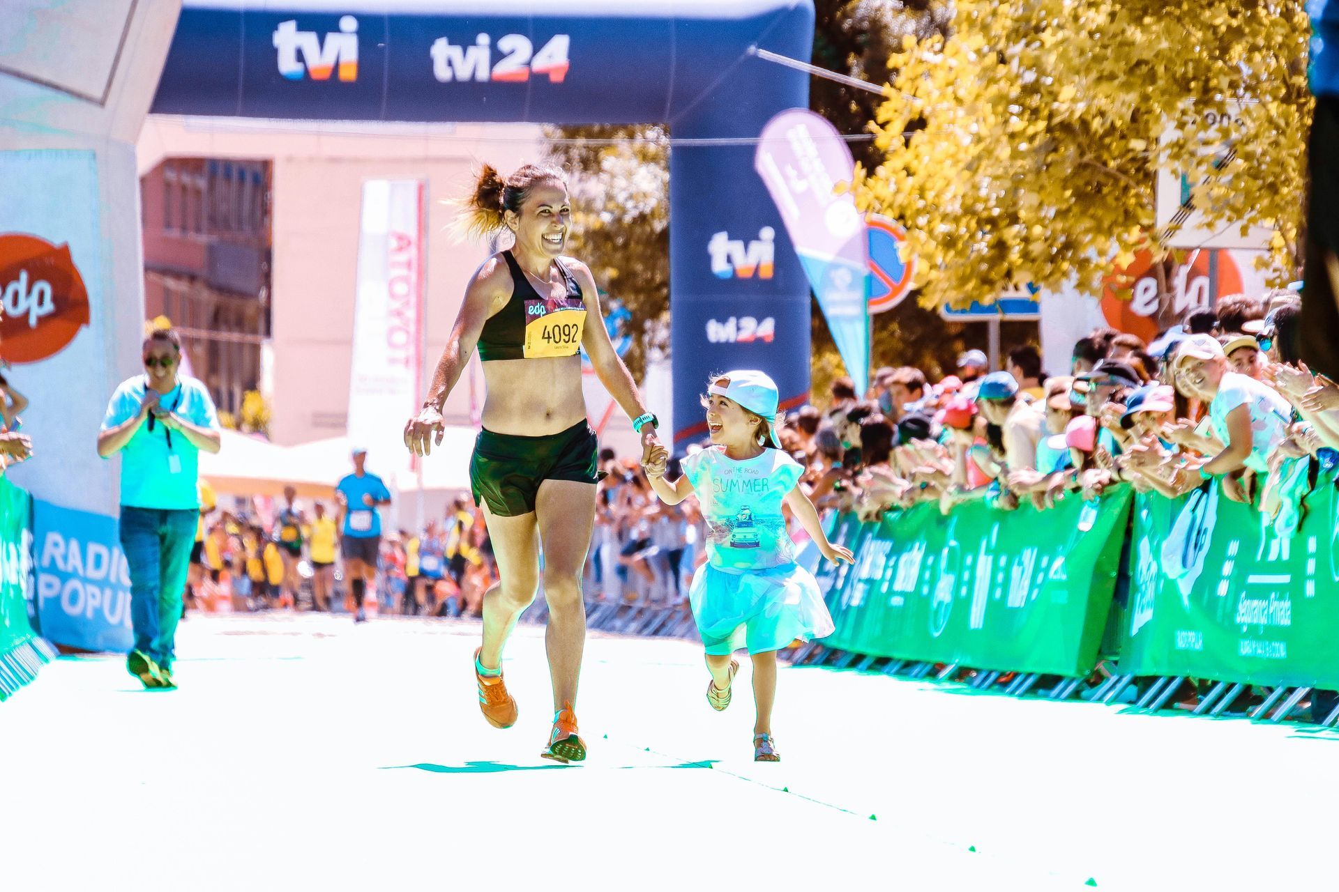 Woman and child run across finish line of a race, hand-in-hand, smiling. Blue, green, and white banners in background.