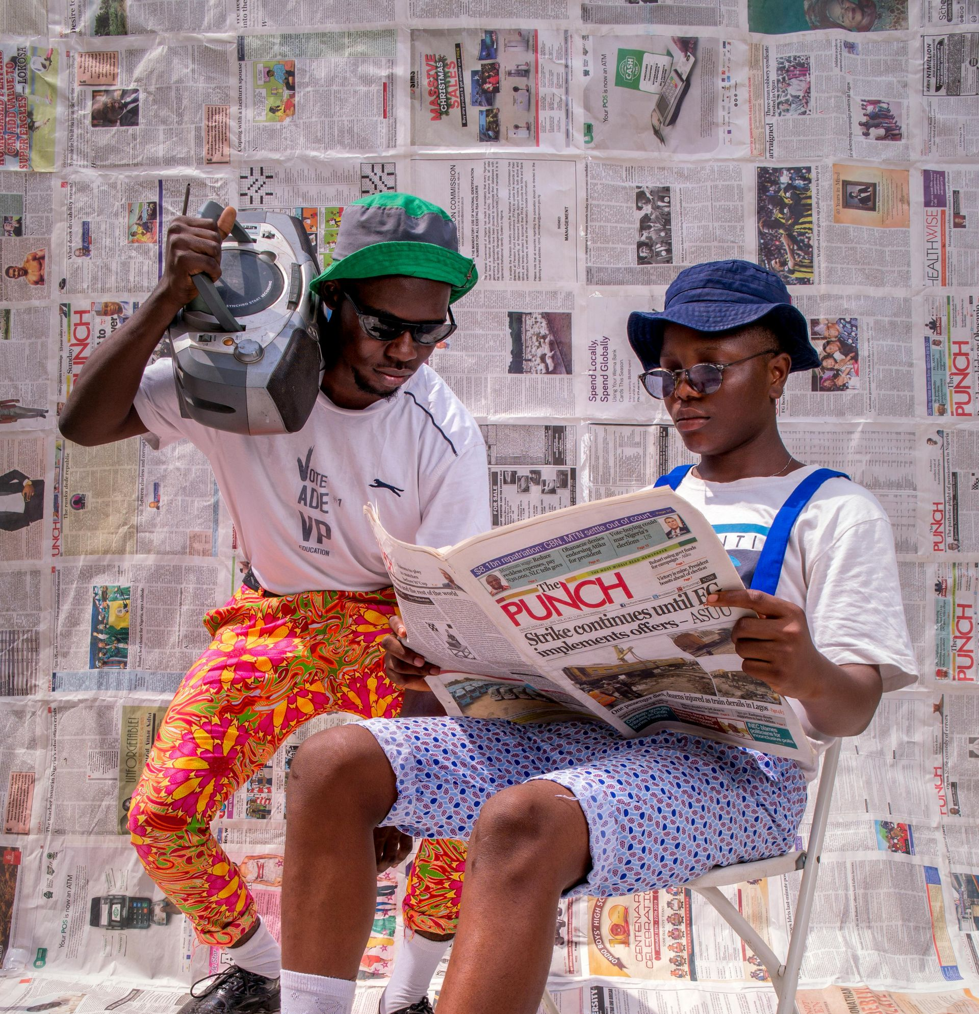 Two people pose in front of a newspaper wall. One holds a boombox, the other reads the 
