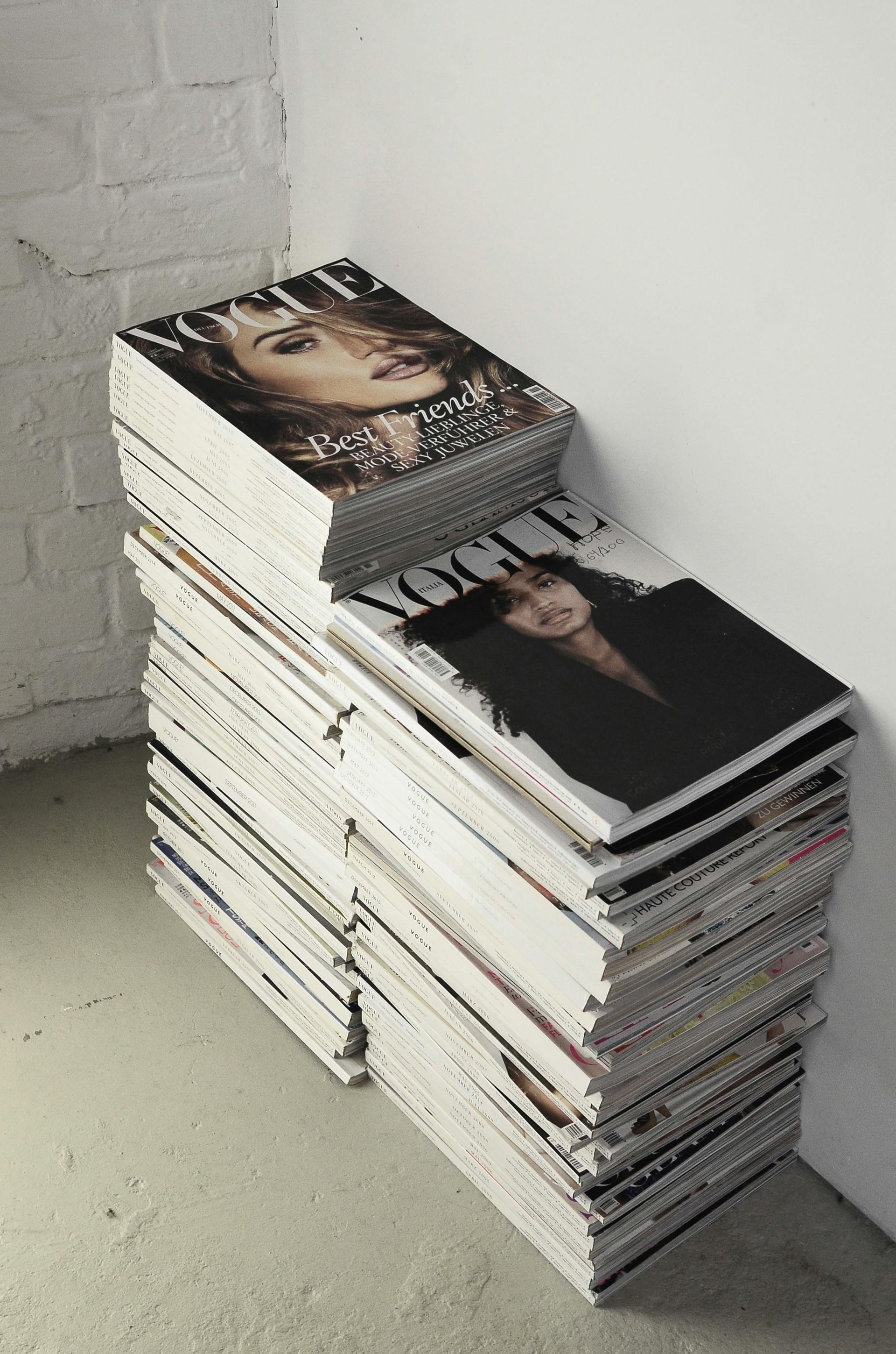 Stacks of fashion magazines piled on a light floor beside a white brick wall and corner.