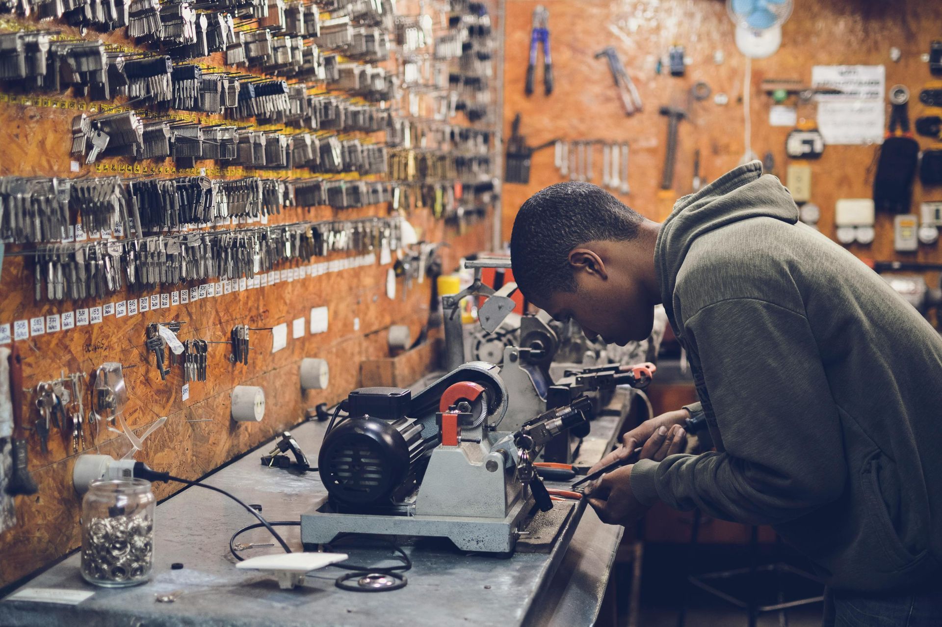 Young Black man using a key-making machine in a cluttered workshop. Many keys line the wall.