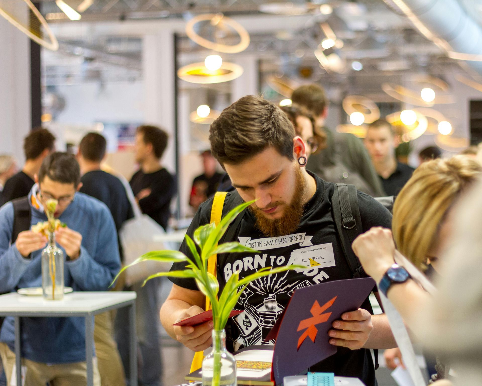Man with beard looks at pamphlet at event, plant in vase on table.