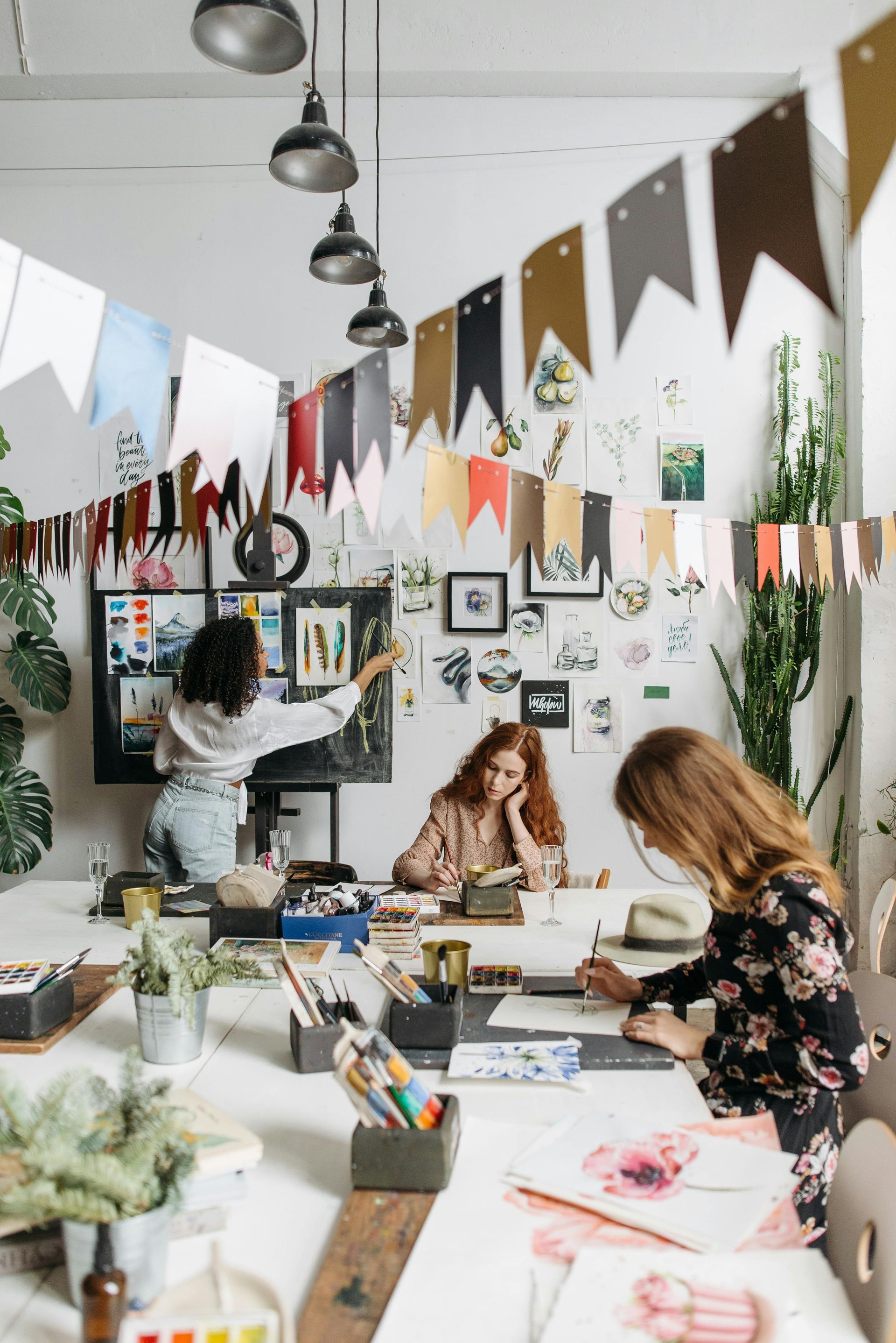 Three women at a creative workspace; one points to a display, others sketch at a white table, decorated with plants and banners.
