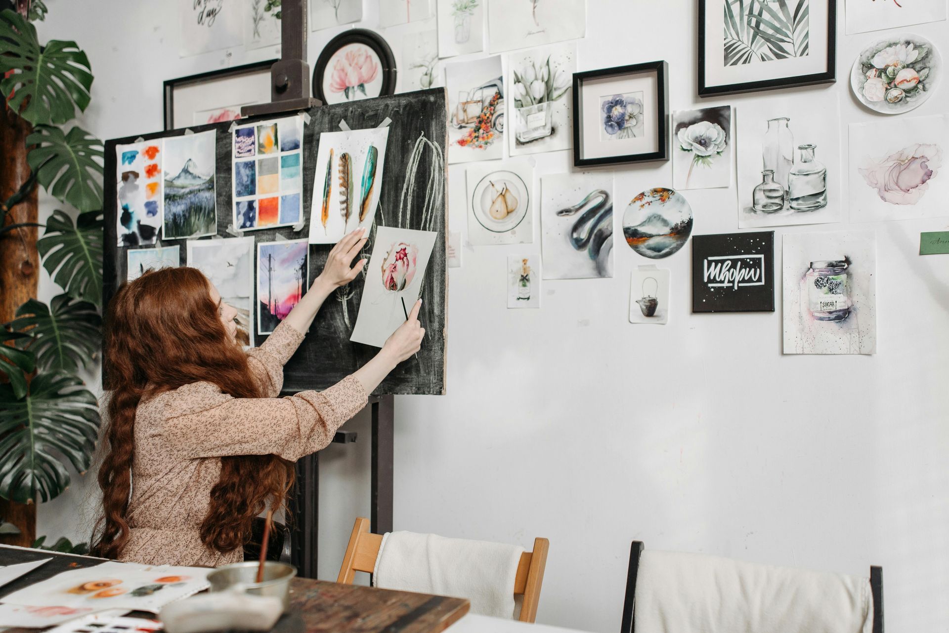 Woman with red hair in art studio, arranging watercolor paintings on a wall, light colors, easel.