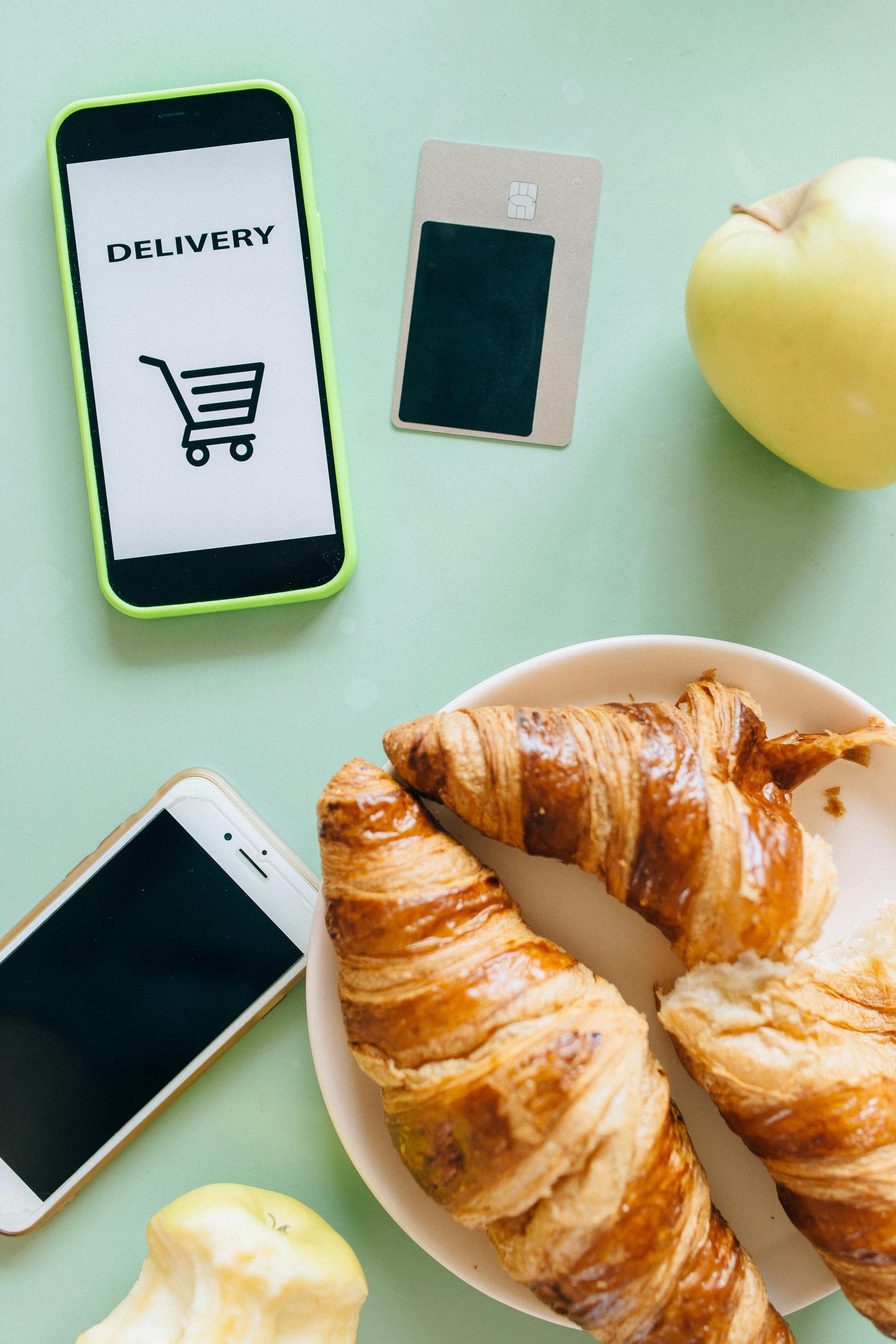 Two phones, a credit card, apple, and croissants. Phone screens show a shopping cart and delivery text.