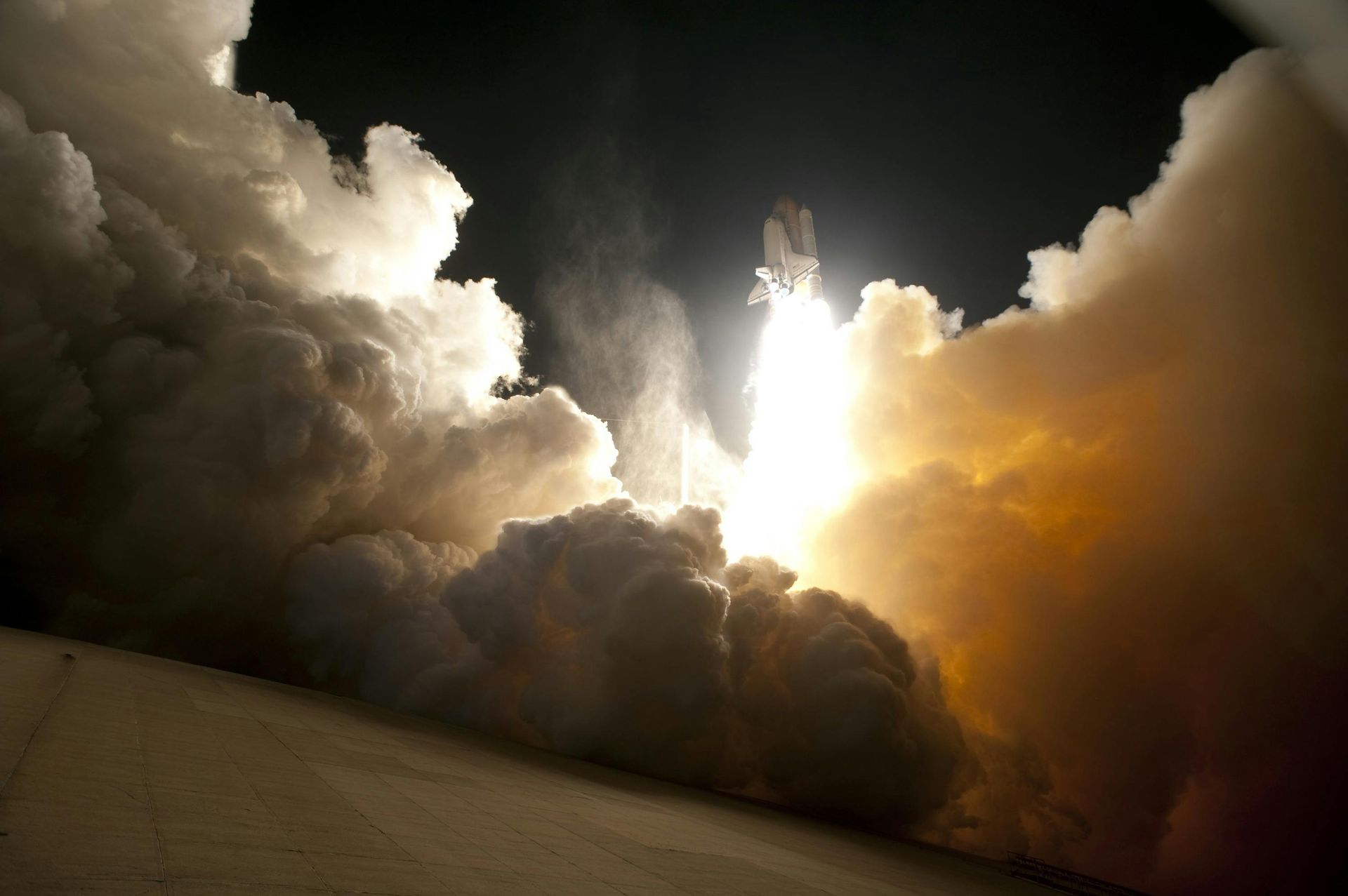 Space shuttle launching at night, bright flames and smoke billowing, dark sky.
