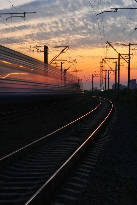 Train speeding along tracks at sunset, motion blur. Orange sky, electrical wires.